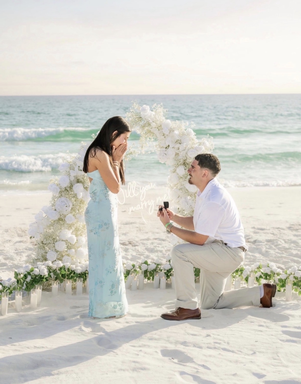 Beachside proposal with white floral arch at sunset